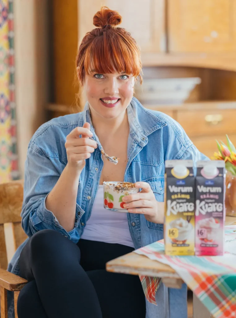Clara Lidström (Underbara Clara) äter Verum Yoghurtkvarg toppad med granola vid köksbord, med förpackningar av Verum i bakgrunden i en ljus hemmamiljö.